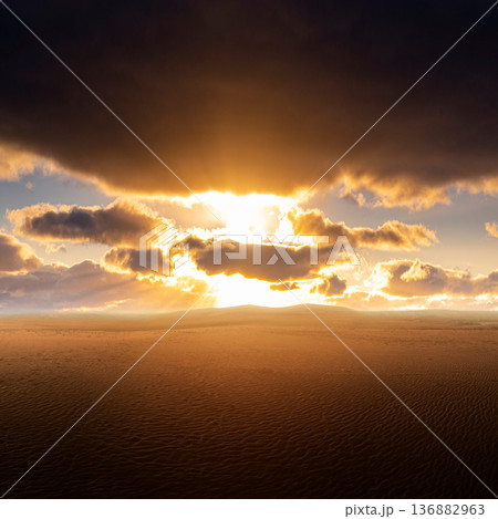 A breathtaking wide view of a vast desert landscape with golden sunlight breaking through dark storm clouds over rolling sand dunes at sunset 136882963