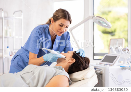 Woman doctor uses hardware ultrasonic to revitalize and tighten facial skin client at clinic Woman doctor uses hardware ultrasonic to revitalize and tighten facial skin client at clinic 136883080