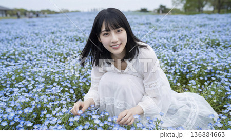 ネモフィラの花畑の中でしゃがみ花を優しく触る白いワンピースの女性 136884663