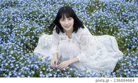 ネモフィラの花畑の中でしゃがみ花を優しく触る白いワンピースの女性 136884664
