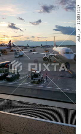 Sunset view from an airport departure lounge through large terminal windows, showing parked airplanes, service vehicles on the apron and passenger reflections in the glass. 136888117