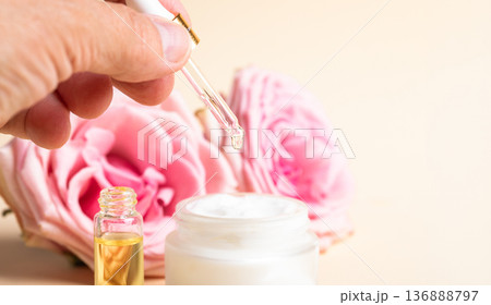 Hand holding dropper with rose essential oil over cream jar and with rose flowers on background 136888797