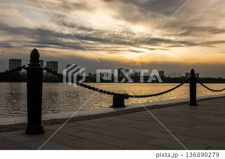 Sunset Over the River With Urban Skyline and Chain-Link Fence Along the Waterfront Promenade in the Evening 136890279