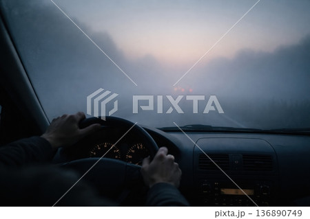 Driving on a Foggy Road at Dawn With Hands on the Steering Wheel and Dim Lights Ahead 136890749
