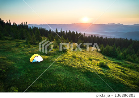 Aerial view of yellow tent on grassy hillside, illuminated by rising sun. Breathtaking panorama of forested mountains and rolling hills under clear sky, creating tranquil and picturesque camping spot. Aerial view of yellow tent on grassy hillside, illuminated by rising sun. Breathtaking panorama of forested mountains and rolling hills under clear sky, creating tranquil and picturesque camping spot. 136890803
