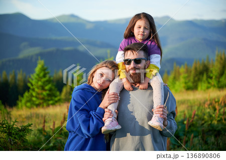 Family stands together in scenic meadow with backdrop of rolling mountains. Father carries his daughter on shoulders, both smiling and enjoying view, while mother stands beside them. Family stands together in scenic meadow with backdrop of rolling mountains. Father carries his daughter on shoulders, both smiling and enjoying view, while mother stands beside them. 136890806