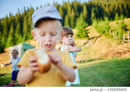 Young girl looks over her shoulder while holding bun. Boy in yellow shirt and cap focuses on his snack in blurred foreground. Another child plays, surrounded by pine trees and rolling hills. Young girl looks over her shoulder while holding bun. Boy in yellow shirt and cap focuses on his snack in blurred foreground. Another child plays, surrounded by pine trees and rolling hills. 136890809
