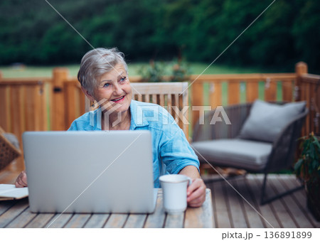 Mature woman sitting on balcony with laptop, working remotely. 136891899