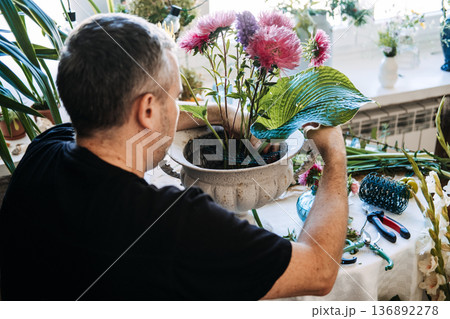 Person arranges aster flowers and green hosta leaf in white decorative bowl. Floral meditation practice, slow arranging flowers, mindful floristry, zen flower therapy, meditative floral design. 136892278