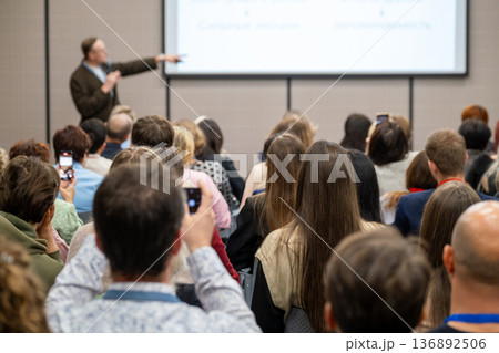 Audience listening to speaker presentation at business conference, attendees using phones to record Audience listening to speaker presentation at business conference, attendees using phones to record 136892506