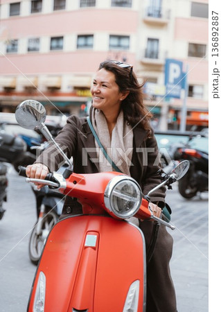 Smiling Woman Riding a Red Scooter in Urban Setting 136892887