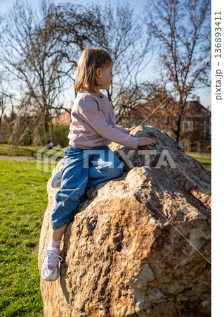 Little girl jumping on decorative stones in city park. Active spring play and outdoor childhood lifestyle. Little girl jumping on decorative stones in city park. Active spring play and outdoor childhood lifestyle. 136893411