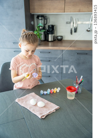 Little girl painting a purple egg at the table. Artistic exploration and childhood development through tradition. 136893415
