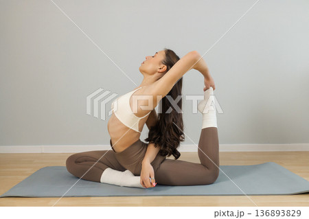 Young flexible woman practicing advanced yoga stretch indoors. Female performing deep backbend pose with leg hold on yoga mat, flexibility training, strength and balance in minimal studio. Young flexible woman practicing advanced yoga stretch indoors. Female performing deep backbend pose with leg hold on yoga mat, flexibility training, strength and balance in minimal studio. 136893829