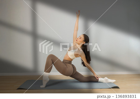 Flexible young woman practicing yoga stretch indoors. Female performing deep lunge side stretch pose on yoga mat, strength, balance and flexibility training in minimal studio with natural light. Flexible young woman practicing yoga stretch indoors. Female performing deep lunge side stretch pose on yoga mat, strength, balance and flexibility training in minimal studio with natural light. 136893831