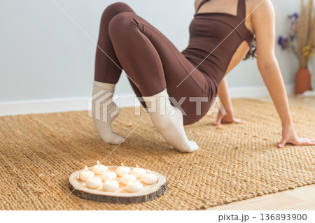 Young woman practicing yoga strength pose indoors. Balance and core training on natural mat, candles nearby, minimal calm interior, wellness, mindfulness and healthy lifestyle concept. Young woman practicing yoga strength pose indoors. Balance and core training on natural mat, candles nearby, minimal calm interior, wellness, mindfulness and healthy lifestyle concept. 136893900