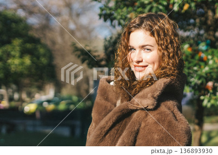 Portrait of young woman outdoors in natural light. Curly hair, warm sunlight, green background, calm expression, lifestyle beauty concept in nature. 136893930