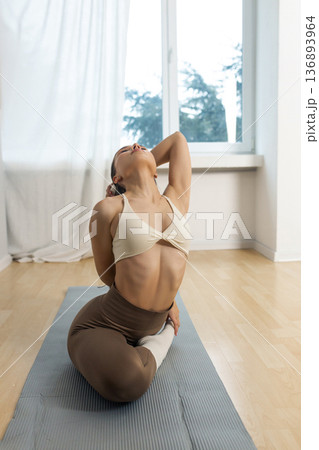 Young flexible woman practicing advanced yoga stretch indoors. Female performing deep backbend pose with leg hold on yoga mat, flexibility training, strength and balance in minimal studio. Young flexible woman practicing advanced yoga stretch indoors. Female performing deep backbend pose with leg hold on yoga mat, flexibility training, strength and balance in minimal studio. 136893964