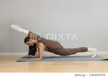 Young athletic woman performing advanced yoga plank variation indoors. Female practicing strength and flexibility exercise on yoga mat, balance control, core training, minimal studio background. 136893970