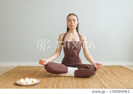 Young woman practicing meditation indoors. Lotus pose on natural mat, closed eyes, burning candles nearby, calm minimal interior, mindfulness and wellness lifestyle concept. 136894061