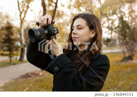 Young woman reviewing photos on her digital camera while standing in a sunny park. Warm autumn light, creative outdoor photography moment, lifestyle scene. 136894151