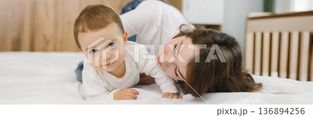 Cozy moment of a young woman and baby relaxing together at home, lying on a soft bed and smiling in natural light. 136894256