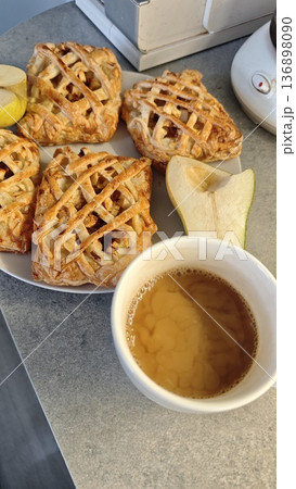 Fresh apple pastry with lattice crust served on plate with pear slice and cup of tea on kitchen table. Concept of cozy breakfast, homemade baking and comfort food lifestyle Fresh apple pastry with lattice crust served on plate with pear slice and cup of tea on kitchen table. Concept of cozy breakfast, homemade baking and comfort food lifestyle 136898090