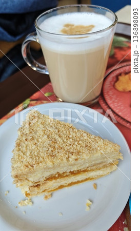 Layered cake slice with cream and fruit filling served on plate with latte in glass cup. Concept of sweet dessert break, cafe atmosphere and cozy lifestyle 136898093