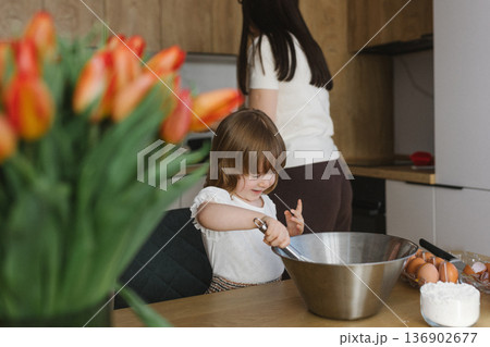 Little girl mixing dough in bowl while mother cooking in background, family baking together at home kitchen 136902677