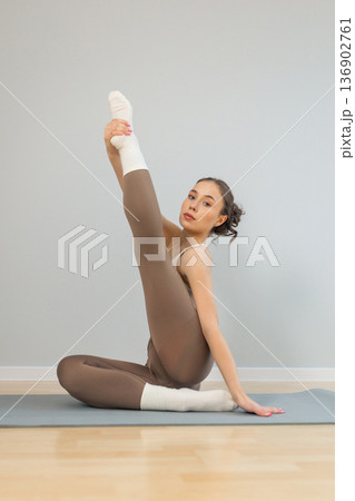 Young woman practicing advanced yoga stretch indoors. Seated pose with raised leg and hand support on yoga mat, flexibility training, balance and body control, minimal studio interior. Young woman practicing advanced yoga stretch indoors. Seated pose with raised leg and hand support on yoga mat, flexibility training, balance and body control, minimal studio interior. 136902761