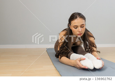 Young woman stretching legs during yoga practice indoors. Forward fold seated pose on mat, flexible athletic body, calm focused expression, minimal studio interior, soft natural light Young woman stretching legs during yoga practice indoors. Forward fold seated pose on mat, flexible athletic body, calm focused expression, minimal studio interior, soft natural light 136902762