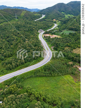 Aerial drone view of Highway 101 winding through lush green mountains between Phrae and Nan, Thailand. A scenic landscape of the road surrounded by tropical forest and a peaceful countryside Aerial drone view of Highway 101 winding through lush green mountains between Phrae and Nan, Thailand. A scenic landscape of the road surrounded by tropical forest and a peaceful countryside 136906642
