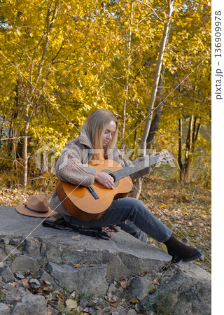 Beautiful blonde girl playing acoustic guitar and singing relaxing enjoying outdoors on sunny autumn day. Hippie woman in boho style learning and practicing playing musical instrument. Fall leaves Beautiful blonde girl playing acoustic guitar and singing relaxing enjoying outdoors on sunny autumn day. Hippie woman in boho style learning and practicing playing musical instrument. Fall leaves 136909978
