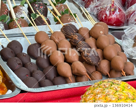 Chocolate coated fruit on sticks at festive market stall. Street dessert culture, indulgence, confectionery craft, and sweet celebration concept. 136910414