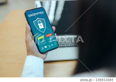 Close up of a person holding a smartphone with an encrypted security interface, featuring shield and lock icons for mobile data protection and digital privacy on blurred office background. Close up of a person holding a smartphone with an encrypted security interface, featuring shield and lock icons for mobile data protection and digital privacy on blurred office background. 136911084