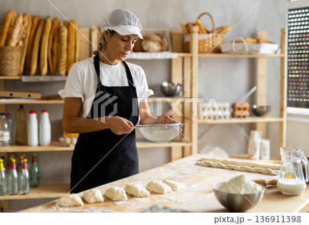 Elderly woman baker sifts flour for dough 136911398