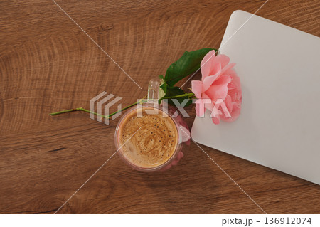 Cup of coffee rests beside a pink rose and a silver notebook on a wooden table Cup of coffee rests beside a pink rose and a silver notebook on a wooden table 136912074