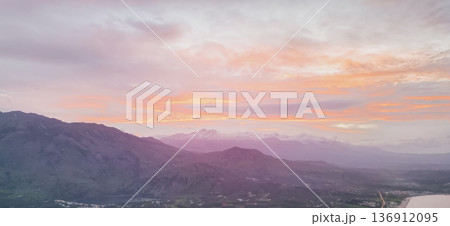 Panoramic sunset view of Crete with shadowed mountain slopes, snow-capped peaks of Lefka Ori, olive valley, coastal village lights, and pastel sky Panoramic sunset view of Crete with shadowed mountain slopes, snow-capped peaks of Lefka Ori, olive valley, coastal village lights, and pastel sky 136912095