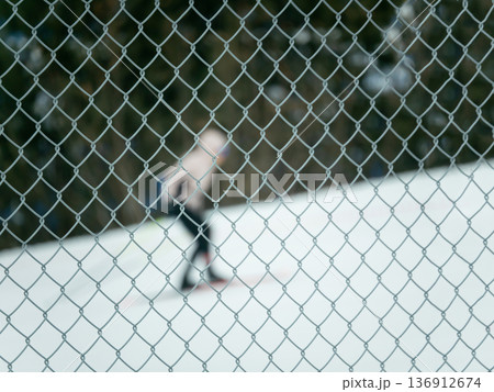 Chain link fence in foreground with blurred person ice skating in background. Winter recreation activity concept for outdoor sports. Chain link fence in foreground with blurred person ice skating in background. Winter recreation activity concept for outdoor sports. 136912674