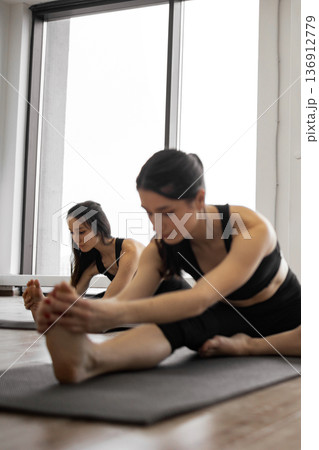 Two women in athletic wear perform a seated forward bend stretching exercise on yoga mats in a bright studio with large windows 136912779