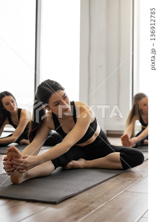 Three women in black activewear practice a seated forward bend on yoga mats in a bright studio with large windows 136912785