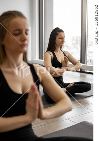 Two women in black activewear practice meditation and mindfulness in a bright yoga studio with large windows Two women in black activewear practice meditation and mindfulness in a bright yoga studio with large windows 136912862