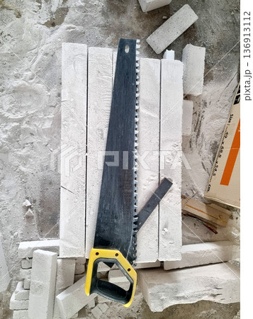 Hand saw with yellow handle resting on stacked aerated concrete blocks at a construction site. High angle view of professional masonry tools and building materials in a dusty environment. 136913112