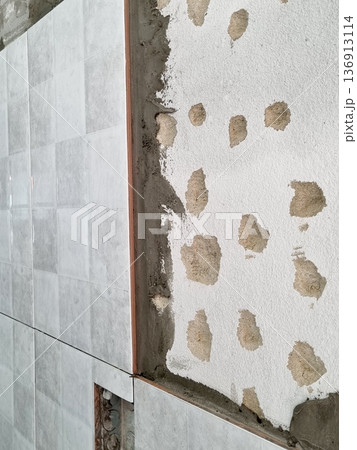 Tiling a wall with grey ceramic tiles during bathroom renovation. Close-up of the installation process showing adhesive mortar on concrete surface. Professional home improvement construction scene. 136913114