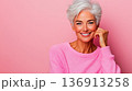 Happy older woman with stylish short hair smiling warmly in a bright pink sweater against a soft pink background during a cheerful indoor moment 136913258
