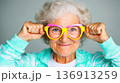 Elderly woman playfully adjusting colorful glasses against a minimalist background in a cheerful indoor setting 136913259