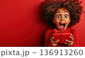 Excitement fills the young girl's face as she holds a beautifully wrapped red gift in front of a vibrant red background during a festive celebration 136913260