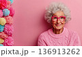 Elderly woman with curly gray hair and pink glasses smiling against a vibrant pink backdrop decorated with colorful pom-poms during a cheerful indoor gathering 136913262
