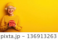 Senior woman in bright yellow sweater and hat holding a red gift box against a vibrant yellow background during a festive celebration 136913263