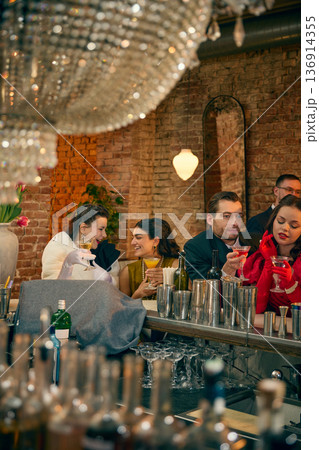 Friends laughing with cocktails at bar counter under crystal chandelier. 136914355